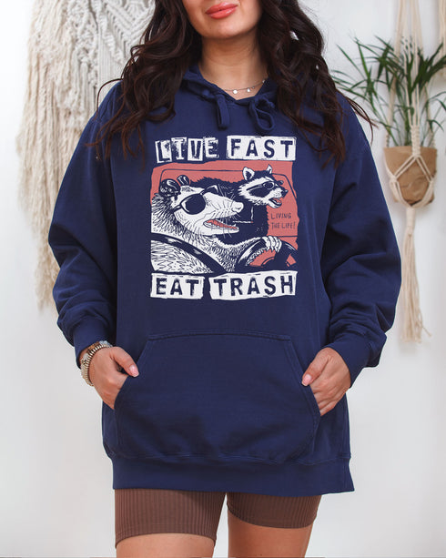 Man wearing a true-navy "Live Fast Eat Trash" hoodie with a retro graphic of a raccoon and opossum driving a car.